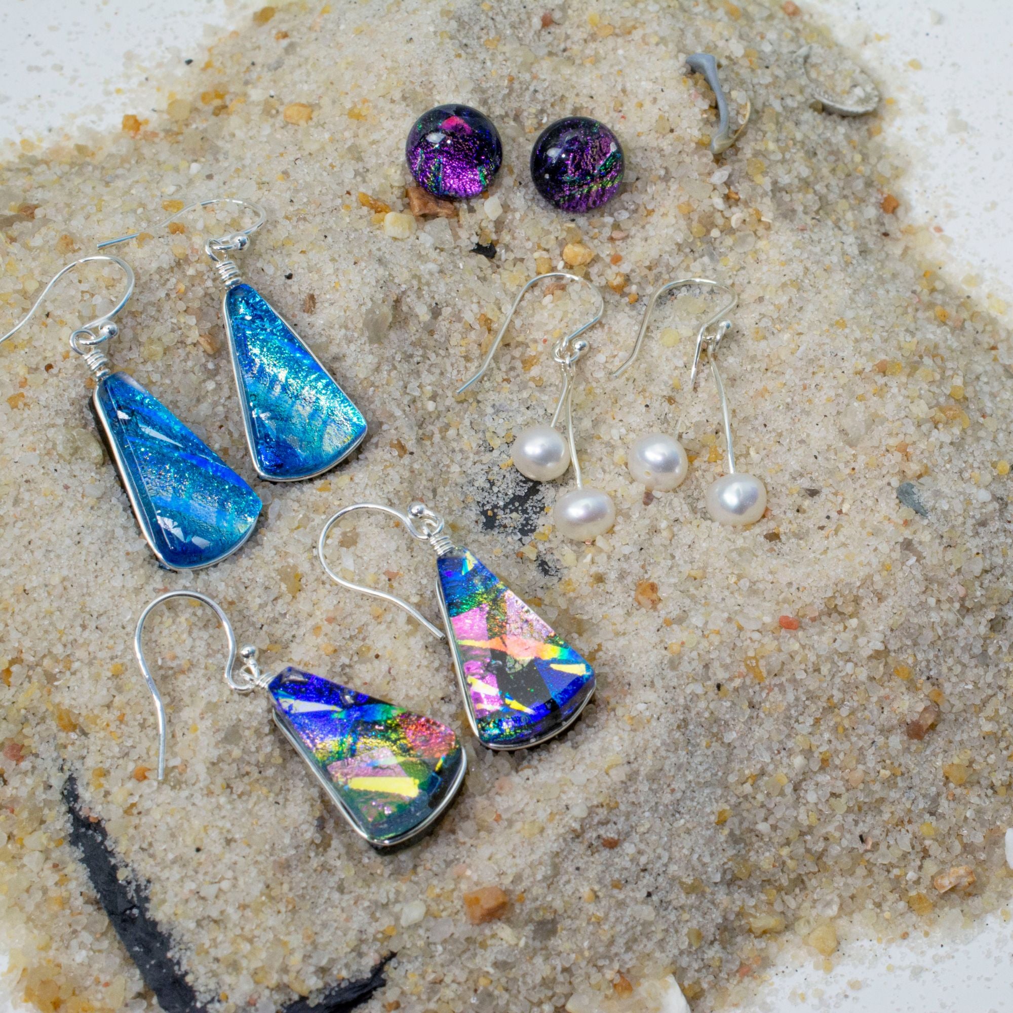 Supernova Earrings displayed on sand with other dichroic glass earrings and pearls.