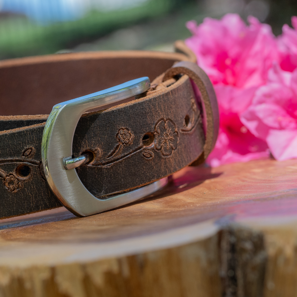 distressed rose leather belt with silver arched buckle on wood round with pink azalea. USA made 