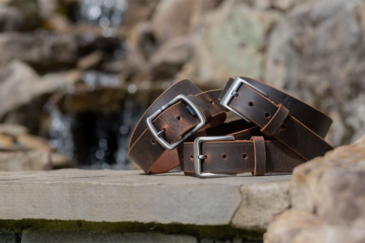 Three brown leather belts with silver buckles on a stone surface with a natural background.